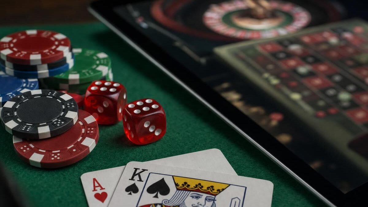Nahaufnahme eines grünen Spieltischs mit Würfeln, Roulettechips und zwei Spielkarten, daneben ein Tablet mit einem unscharfen Spieltisch auf dem Bildschirm, gedämpftes Licht