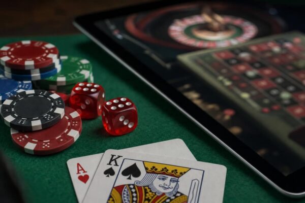 Nahaufnahme eines grünen Spieltischs mit Würfeln, Roulettechips und zwei Spielkarten, daneben ein Tablet mit einem unscharfen Spieltisch auf dem Bildschirm, gedämpftes Licht